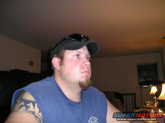 Man wearing a blue t-shirt, black baseball cap, and sunglasses, looking to the right. He has a goatee and tribal tattoos on his left arm