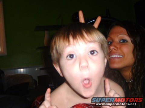 Child with fingers on head like bunny ears, woman smiling behind. Supermotors logo at bottom right