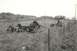 1967 Ford F-100 - A damaged truck chassis lies on its side in a grassy field, with several people gathered near another vehicle by a fence.