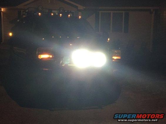 Dark-colored Ford Excursion front view at night, headlights on, roof rack with lights, in front of a house