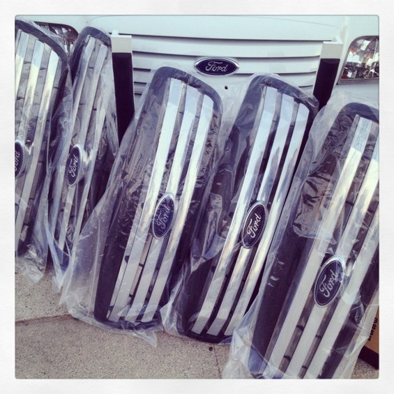 Several new Ford grilles in plastic packaging, stacked in front of a white vehicle