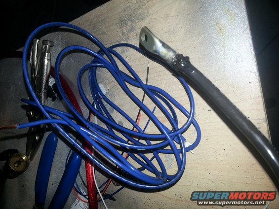 Blue wires, red wire, and tools like pliers and a vise grip are on a wooden surface next to a thick black cable with a metal connector