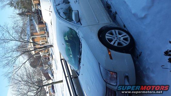 White sedan parked in snow, front quarter view. Houses and bare trees in background