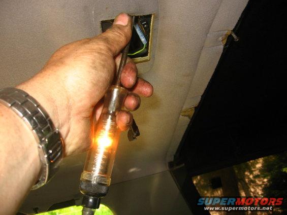 Hand holding a circuit tester to a wire in the ceiling of a vehicle