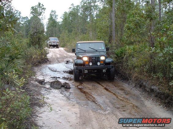 Green Jeep Wrangler driving on a muddy dirt road, front view. Another SUV is visible in the background