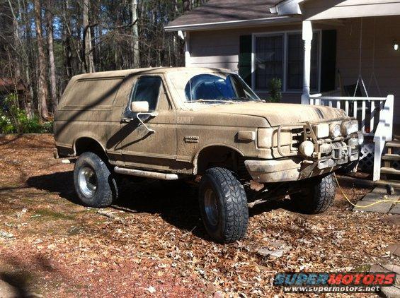 1988 Ford Bronco covered in mud, parked in a yard with a house in the background