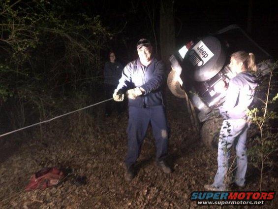 People pulling a Jeep out of the woods with a rope. One person is holding the rope, another is standing near the Jeep