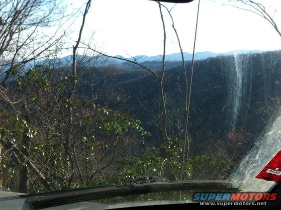 Dashboard and windshield of a vehicle looking out at a forest with mountains in the distance. A red sticker is visible on the windshield