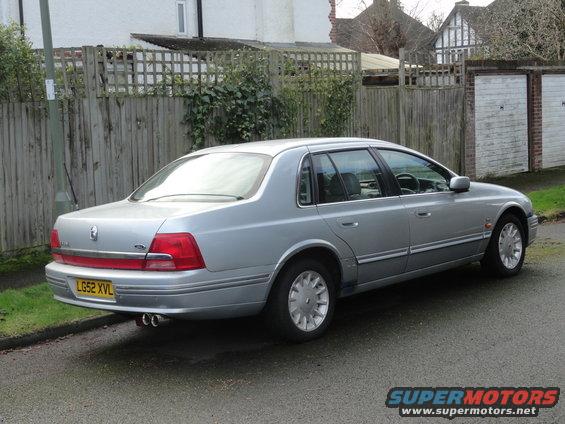 Silver sedan parked on a street, rear quarter view. The car has a yellow license plate and dual exhaust pipes