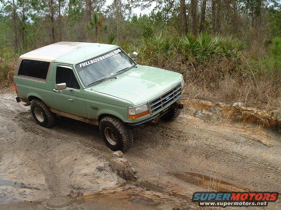 Green 1995 Ford Bronco driving on a dirt trail with trees in the background. It has a banner on the windshield that says FULLSIZEBRONCO.COM