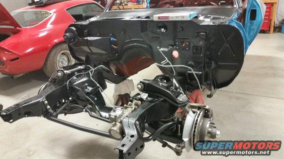 Car chassis and engine bay disassembled, with a red 1969 Chevrolet Camaro in the background
