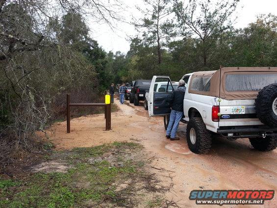 White 1994 Ford Bronco with tan soft top, rear view, on a dirt road with other vehicles