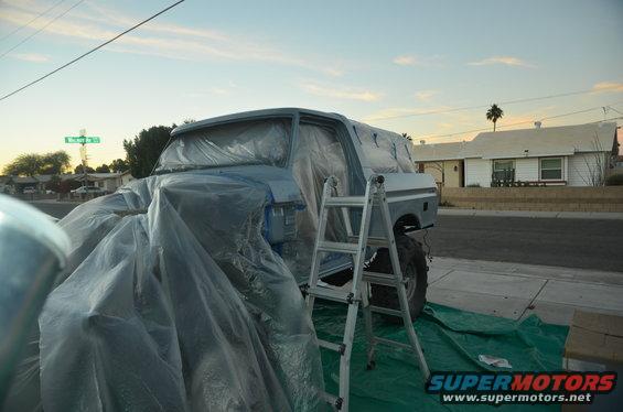 1979 Ford Bronco covered in plastic sheeting with a ladder next to it, parked on a street