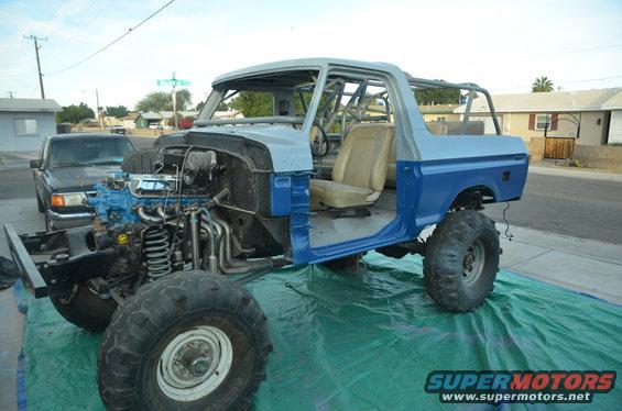 Blue and gray Ford Bronco body with exposed engine and roll cage, sitting on a green tarp. A black pickup truck is parked behind it