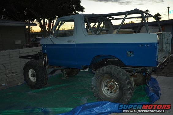 Two-tone blue and white Ford Bronco with a roll cage and large off-road tires, jacked up