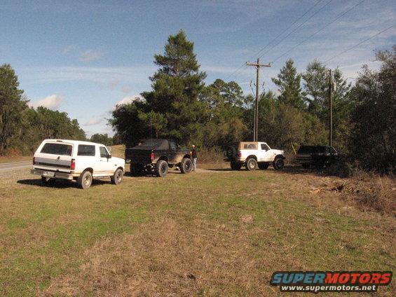 Four SUVs parked in a grassy field with trees and power lines in the background. The white SUV on the left is a 1987 Ford Bronco, viewed from the rear-side