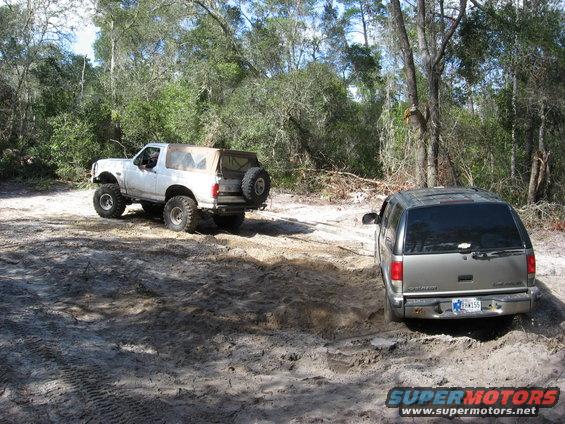 White Bronco and silver SUV driving on dirt path in woods