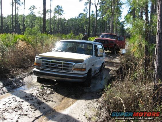 White 1992 Ford Bronco and red Ford truck on muddy forest trail