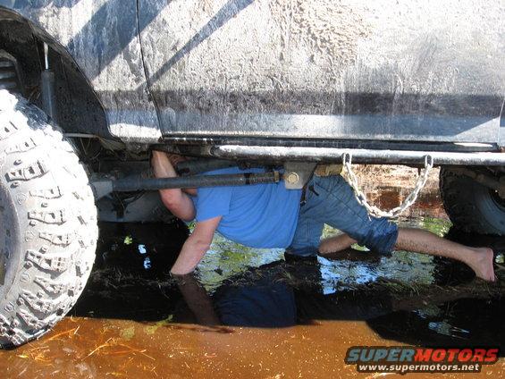 guy working under a muddy SUV in shallow water, big tire visible