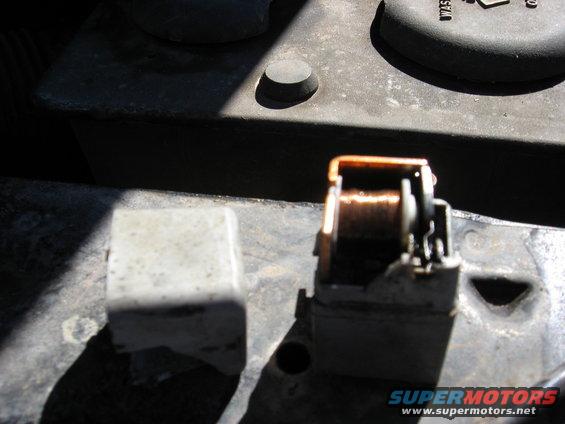 Automotive relay with copper coil, next to a grey plastic cover, on a dark metal surface