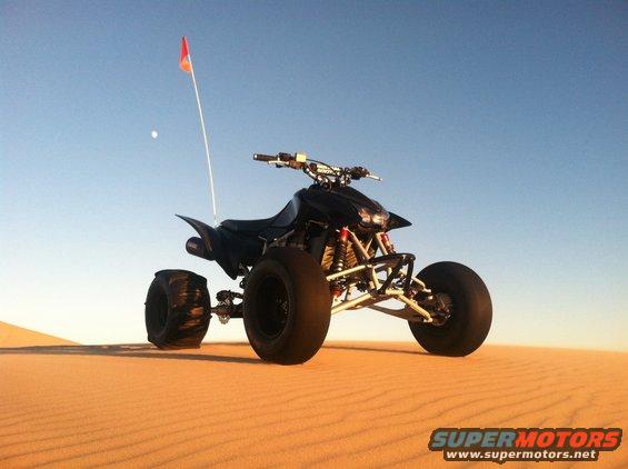 Black ATV with a red flag on a sand dune under a clear blue sky
