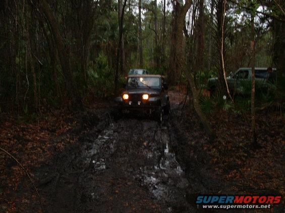 Dark Jeep Wrangler driving through a muddy forest path, with another vehicle visible in the background