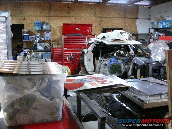Car body in a workshop, engine exposed, parts scattered around. A red toolbox is in the background