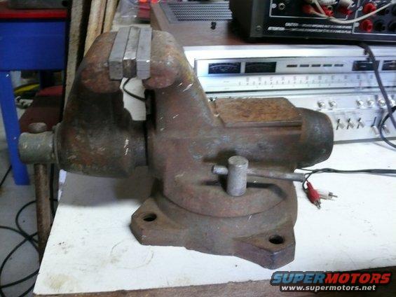 Rusty bench vise on a white surface with audio cables connected to electronic equipment in the background