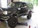 1988 Ford Bronco - A black Ford Bronco is shown with its front wheel raised on a creeper, revealing its suspension and custom wheels.