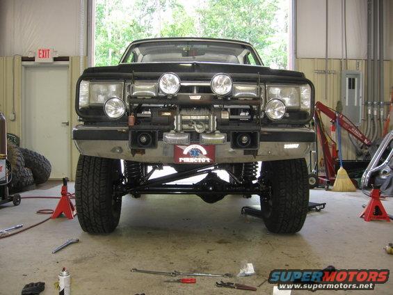 Front view of a modified Ford Bronco, lifted with large tires and a winch mounted on the bumper. The vehicle is in a garage with tools on the floor
