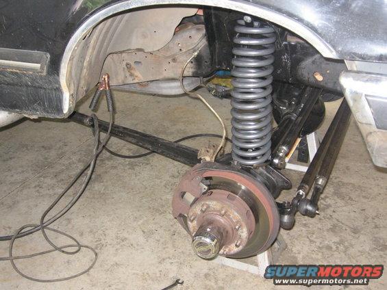 Vehicle front suspension with large coil spring, brake rotor, and tie rod assembly. Black body panel visible