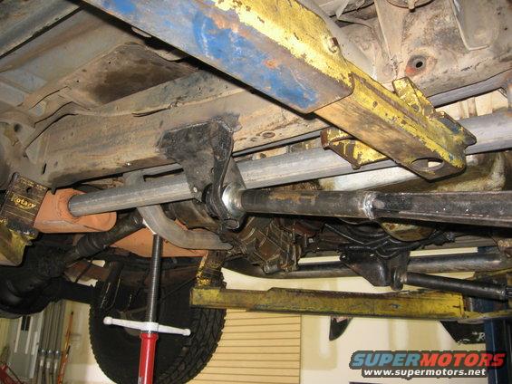 Underbody view of a vehicle on a lift, showing suspension, exhaust, and drivetrain components. SuperMotors logo visible