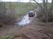 1976 Ford F-100 - A dark SUV navigates a muddy, waterlogged dirt track surrounded by vegetation.