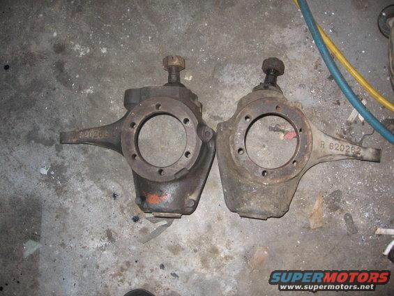 Two vehicle steering knuckles, one darker than the other, resting on a concrete floor. Part numbers are visible
