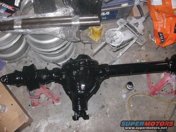 black painted rear axle assembly with driveshaft, sitting on red jack stands, with spare wheels and a bag of pet food in background