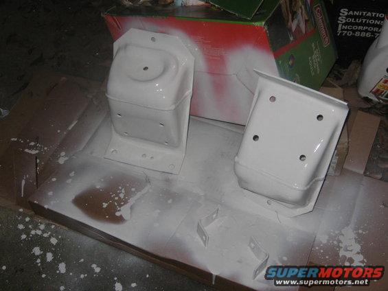 Two white painted metal brackets on a cardboard surface with overspray