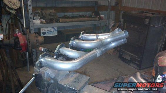 custom exhaust headers clamped in a vise, with a vehicle axle and shelves in the background