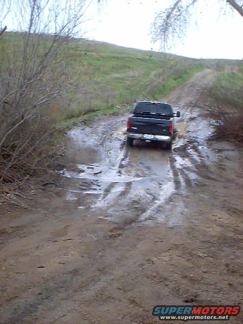 Black pickup truck driving through a muddy, wet dirt road with puddles