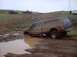 1976 Ford F-100 - A pickup truck is stuck in a mud pit with other vehicles visible on a nearby hill.