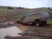 1976 Ford F-100 - A pickup truck is stuck deep in the mud, with other vehicles visible on a nearby hill.