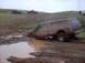 1976 Ford F-100 - A muddy pickup truck with a camper shell is stuck in a muddy rut, with other vehicles in the background.