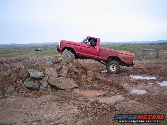 Red 1976 Ford F-100 climbing over a pile of rocks on a muddy off-road course. Another vehicle is visible in the background