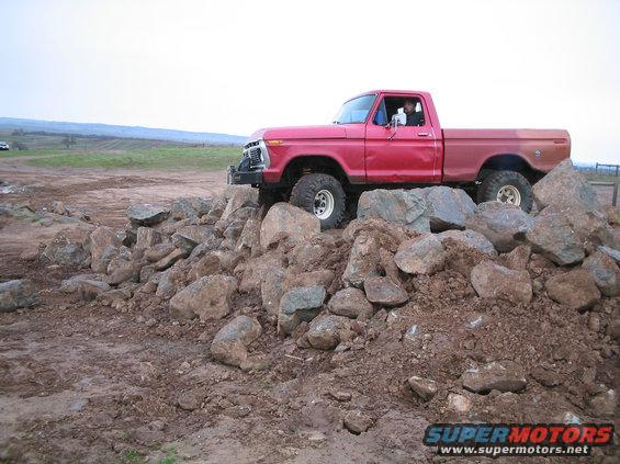 Red pickup truck driving over a pile of rocks. The truck has large tires and a winch on the front
