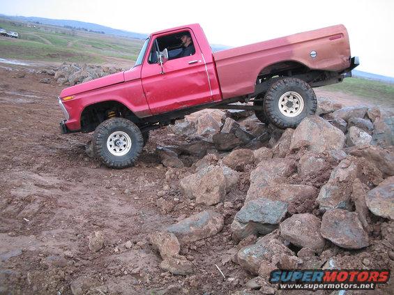 Red pickup truck with lifted suspension driving over rocks. Visible text: SUPERMOTORS.NET