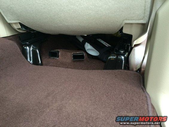 Under seat storage compartment with brown carpet and black metal brackets. A white plastic piece is visible