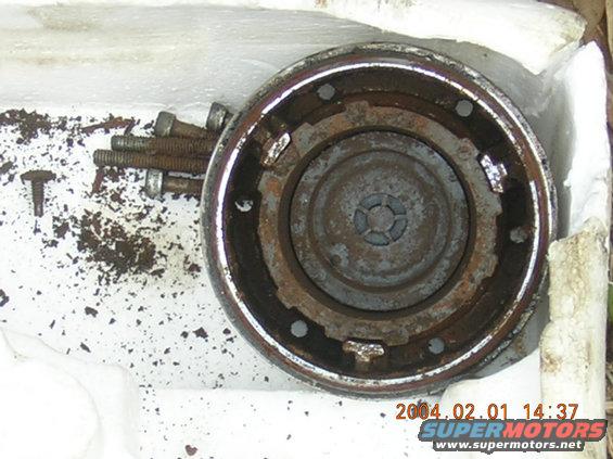 Rusted circular metal part with four bolts and debris on a white surface