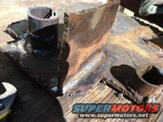 Rusty metal bracket with visible welds and a pipe section attached, resting on a wooden surface