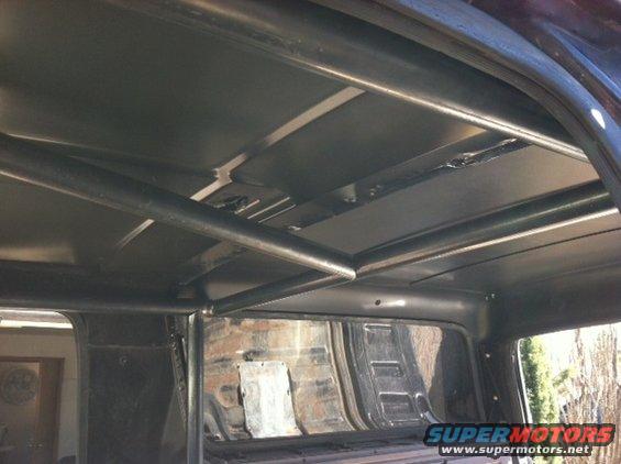 Interior view of a vehicle with a custom roll cage and metal ceiling panels. Visible brickwork and a window are in the background