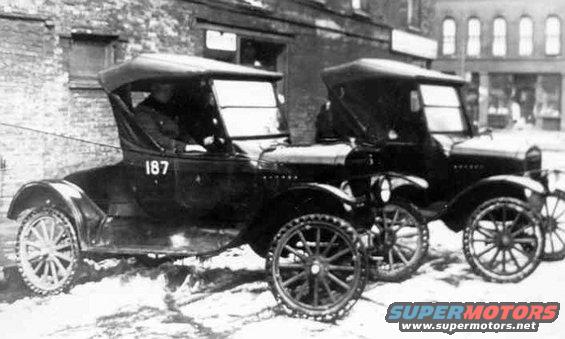Two early 1900s cars parked side-by-side in the snow. One has '187' on its door
