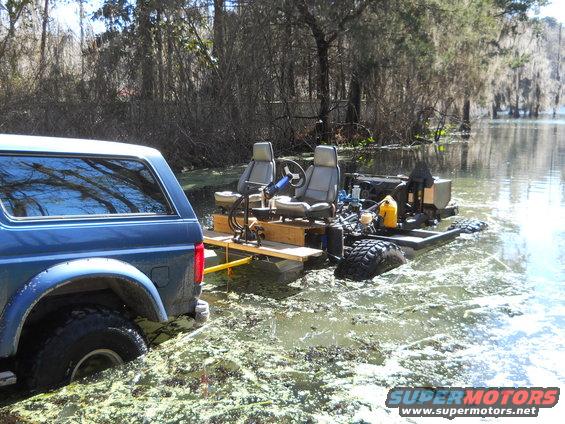 Blue 1992 Ford Bronco partially submerged in murky water, attached to a homemade amphibious vehicle with two seats